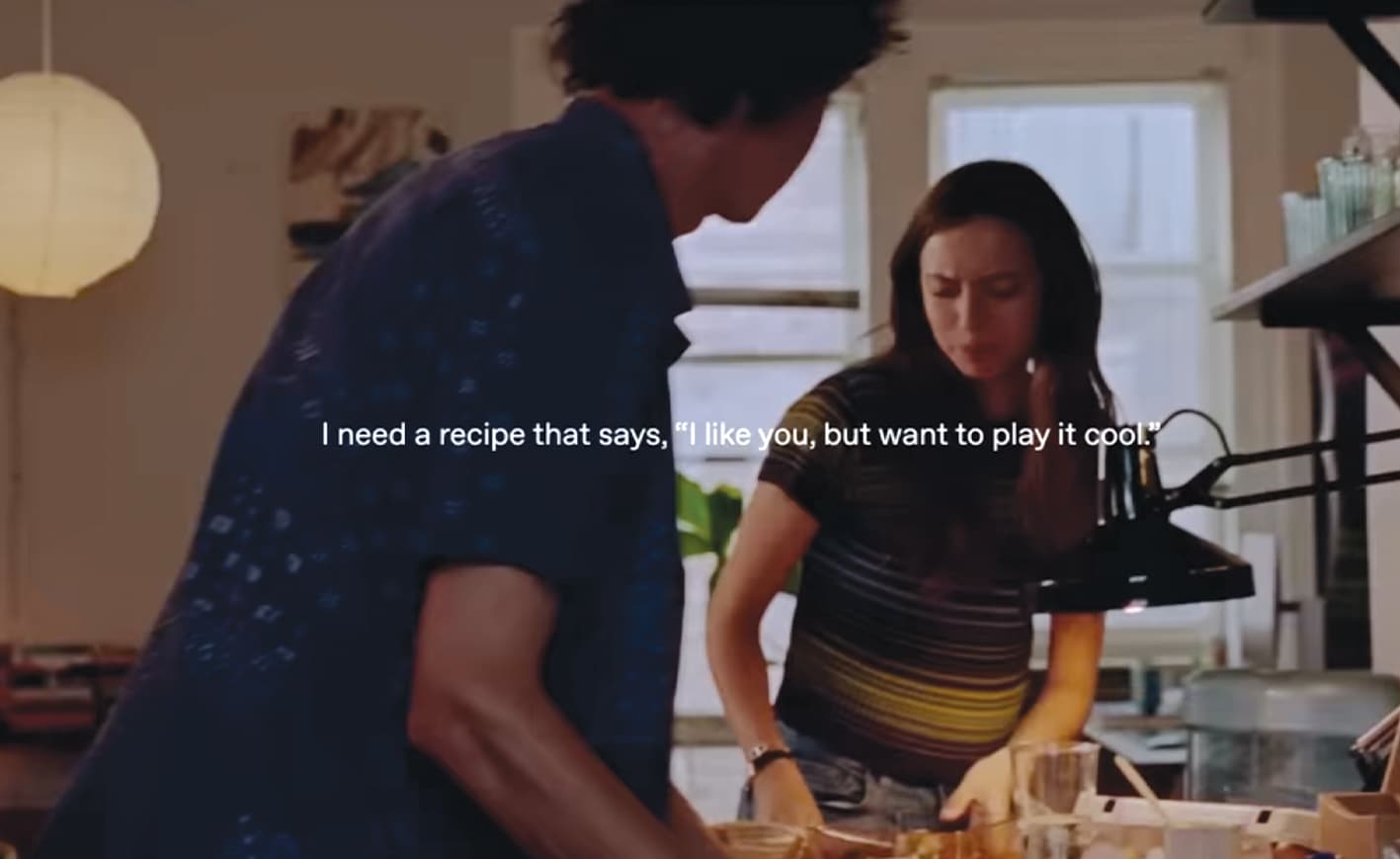 A photo still of a video in which a man and woman are together in a kitchen. Overlaying the image are the words "I need a recipe that says, 'I like you, but want to play it cool.'"